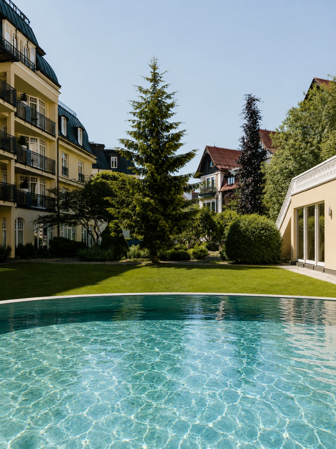 Thermal pool in the garden of a Belle Époque spa hotel in Mariánské Lázně, Czech Republic — one of the quieter European spa towns — with yellow-and-green hotel facades and pine trees reflected in the still water