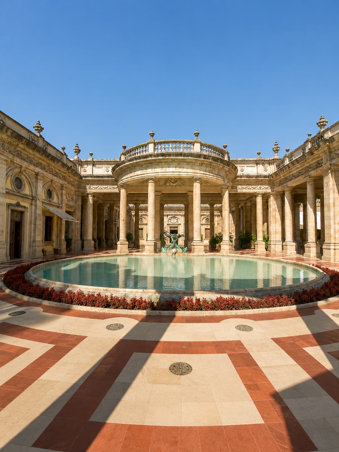 The neoclassical colonnaded courtyard of Terme Tettuccio in Montecatini Terme, Italy — a UNESCO Great Spa Town of Europe — with a circular fountain pool, marble floors, red flower borders, and ornate arched loggia