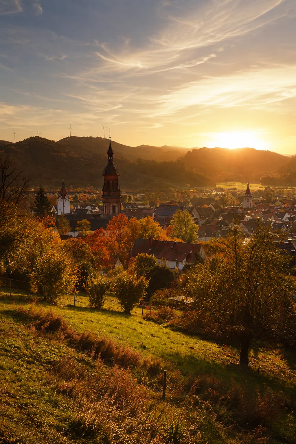 A Black Forest village at golden hour in autumn — church tower and rooftops surrounded by amber trees, sun low on the hills behind