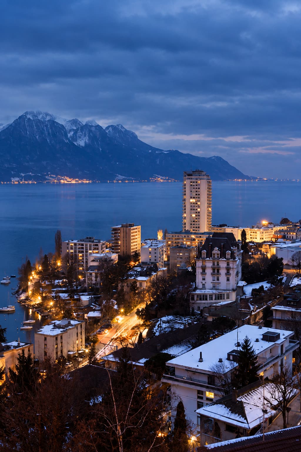 Montreux city at blue hour in winter, amber street lights reflected in Lake Geneva with snow-capped Alps rising across the water under a deep blue sky