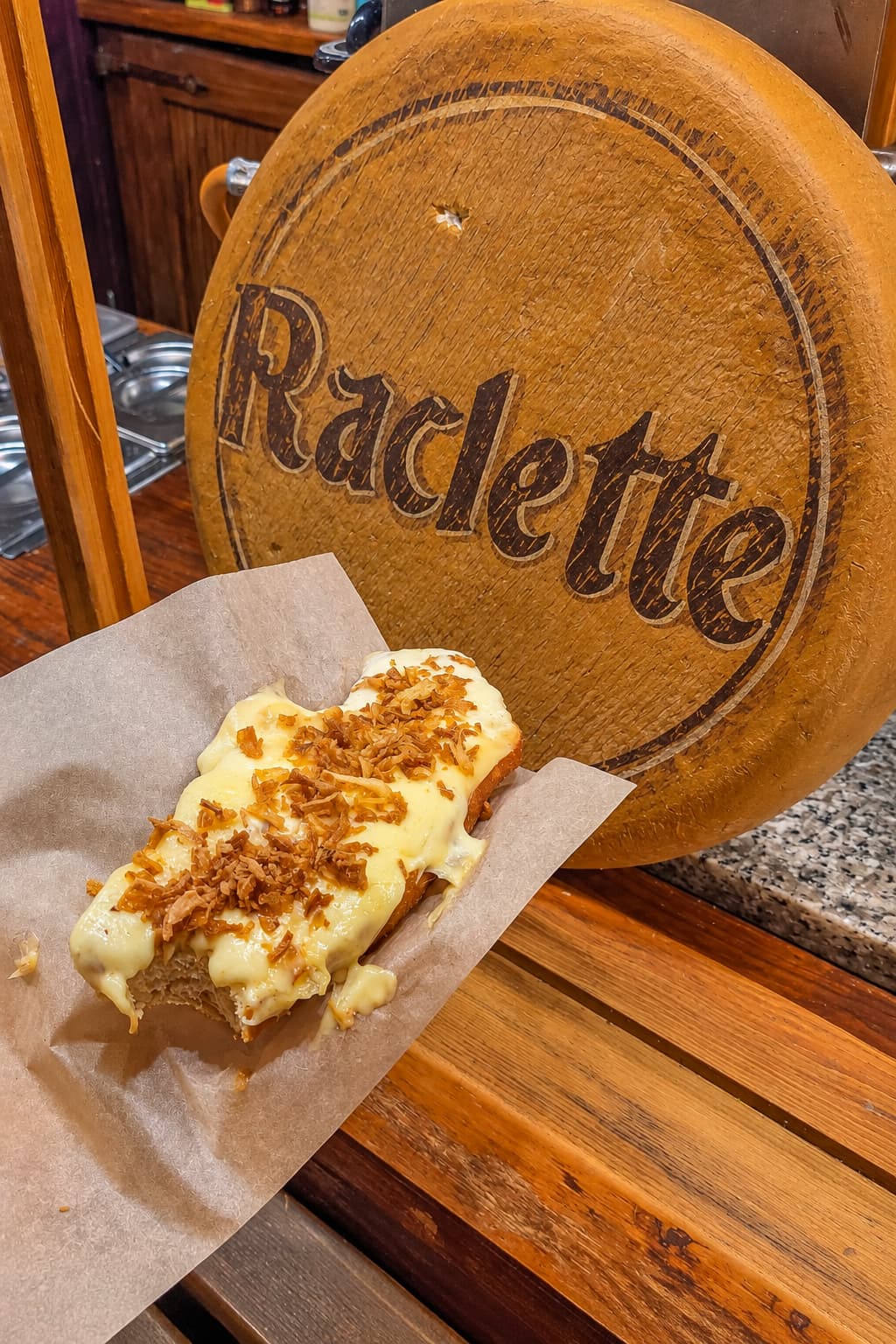 Raclette on bread with caramelised onions served on parchment paper beside a round wooden Raclette sign at a Swiss Christmas market stall