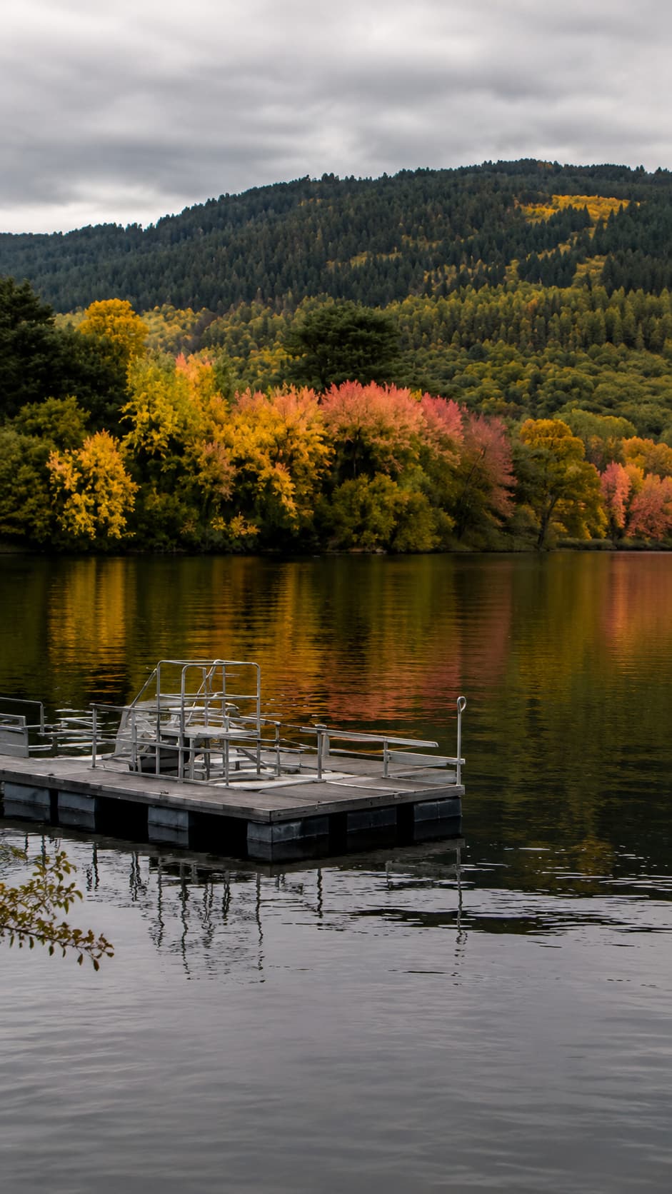 A floating dock on a Scottish loch in autumn, vivid orange and red tree reflections in still grey water under an overcast sky