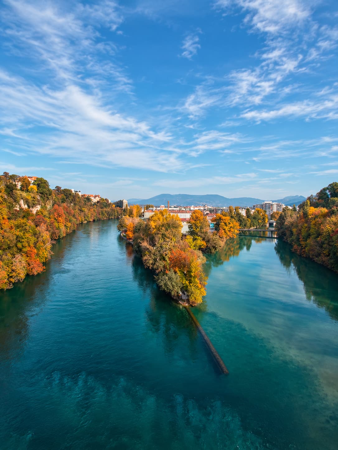 Aerial view of the confluence of two rivers in central Switzerland in autumn, with autumn-coloured trees lining the turquoise water and a town and mountains visible in the distance