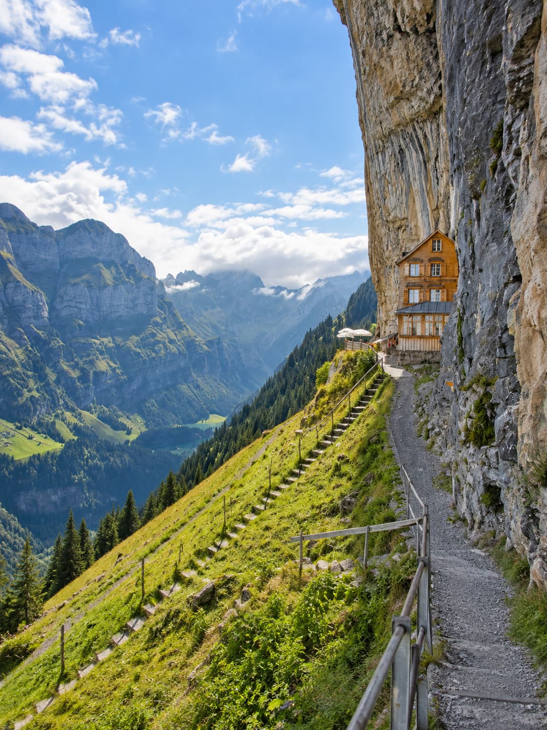 Narrow mountain path carved into a cliff face in Appenzell, Switzerland, leading past the Äscher guesthouse perched under an overhanging rock, with a deep green valley and dramatic mountain walls stretching below