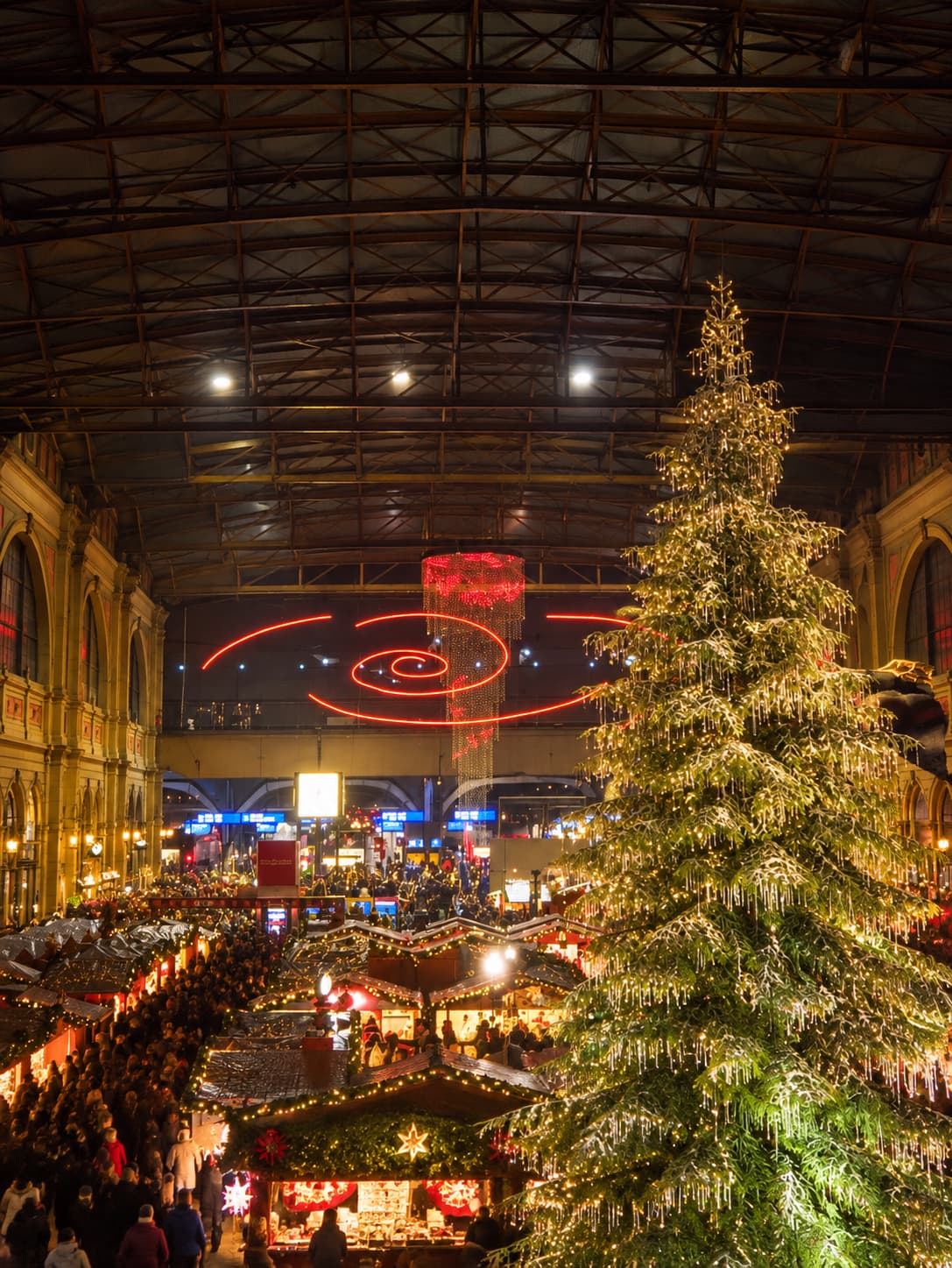 Zurich Hauptbahnhof Christmas market interior — illuminated tree and market stalls inside the grand station hall, with a red chandelier installation hanging from the arched roof above