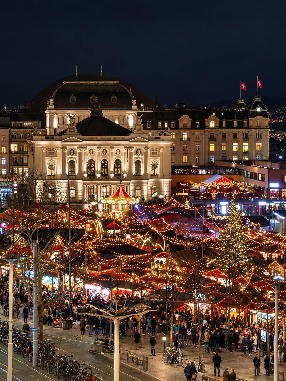 Aerial night view of Zurich's Wienachtsdorf Christmas market at Sechseläutenplatz — a sea of golden lights and red-roofed market stalls with the illuminated Opera House behind and Swiss flag visible on the rooftop