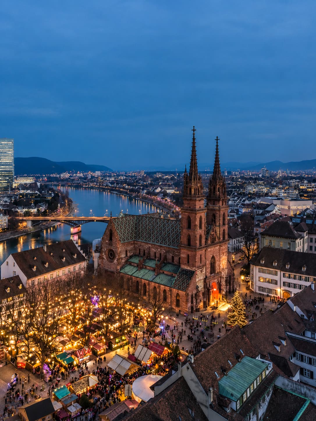 Aerial dusk view of Basel's Christmas market at Münsterplatz — golden market stall lights cluster around the red-sandstone Basel Minster cathedral with twin spires, the Rhine river and city beyond glowing in deep blue twilight
