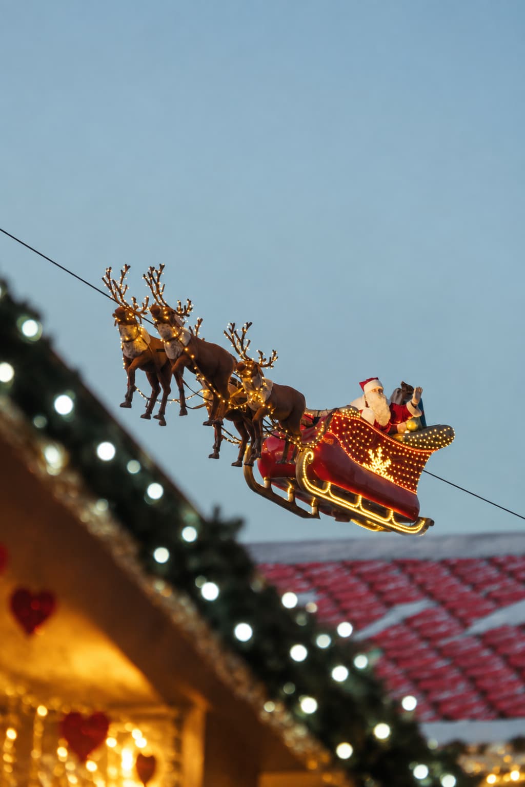 An illuminated Santa Claus sleigh with reindeer gliding on a wire above the Montreux Christmas market rooftops at dusk, against a pale blue winter sky