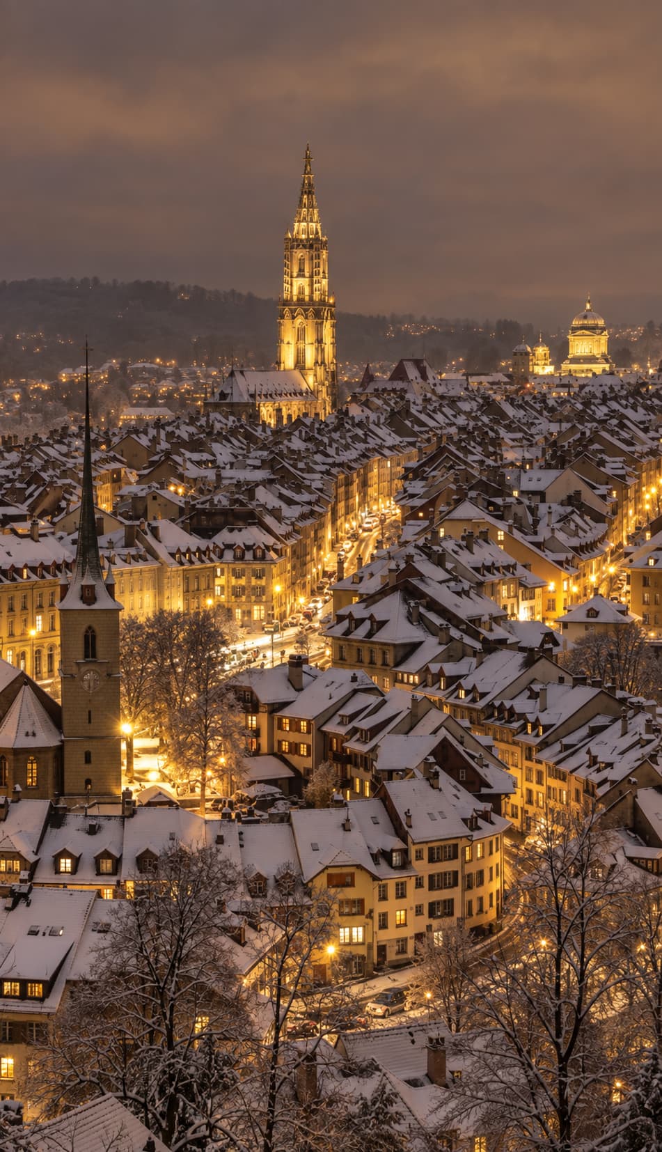 Bern old town in winter at night — snow-covered rooftops stretch across the frame, the illuminated spire of Bern Minster rises in the centre, and amber streetlights glow along the historic streets below a dark cloudy sky