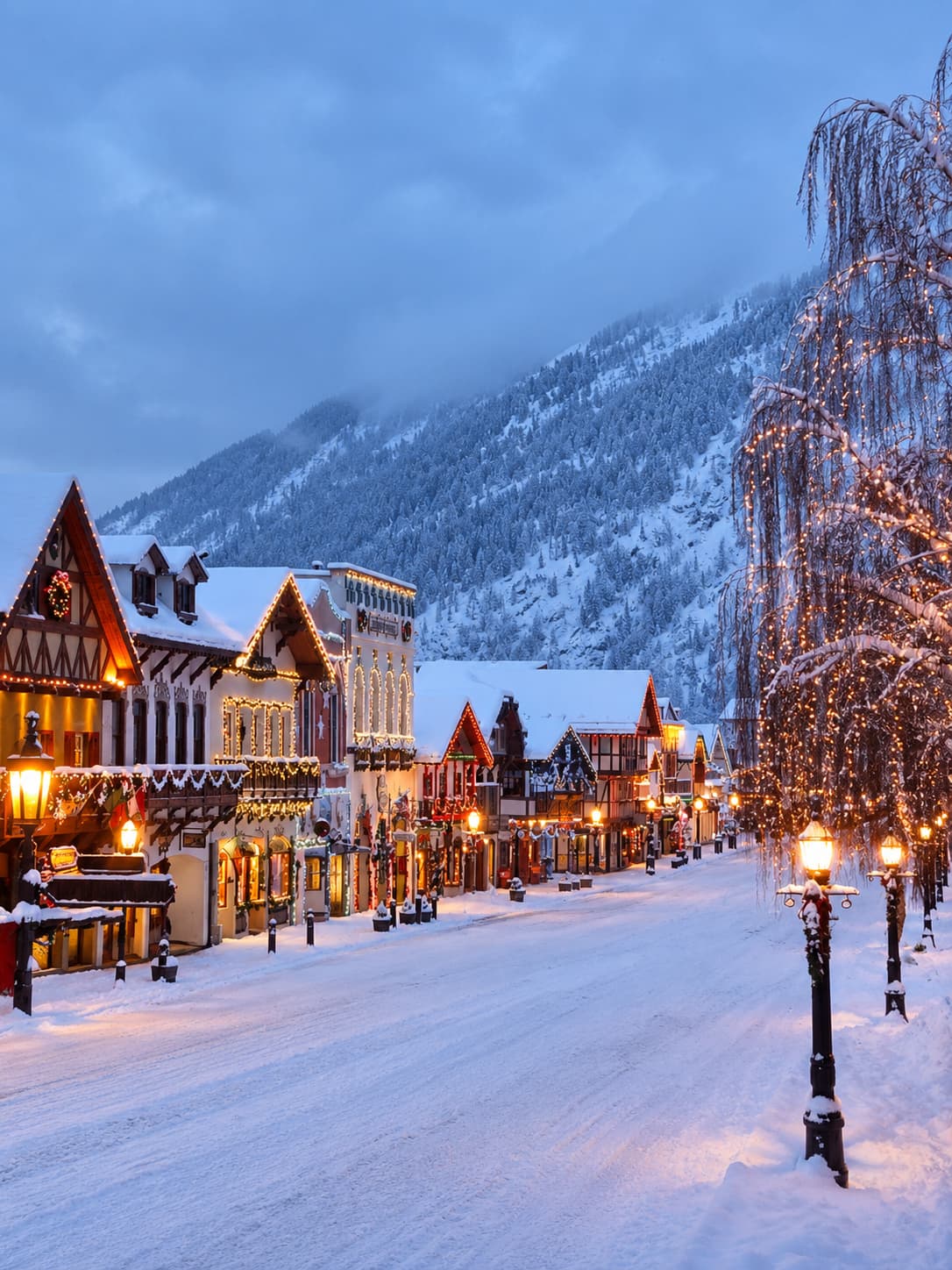 A snow-covered alpine village street at blue-hour dusk — timber-framed buildings with warm Christmas lights line both sides of the empty road, with a snow-dusted forested mountain rising directly behind the village