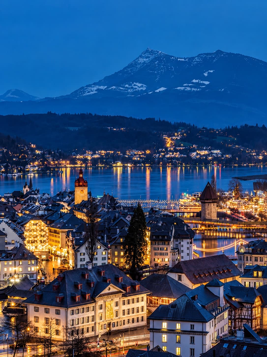 Lucerne old town at blue hour — the illuminated Chapel Bridge and its water tower are visible crossing the river, golden lights reflect off Lake Lucerne, and a snow-capped Alpine peak rises against the deep blue night sky behind the city