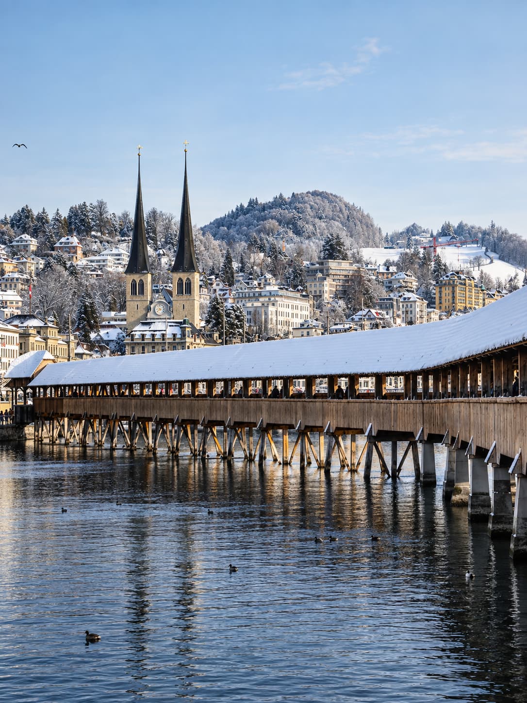 Lucerne's Chapel Bridge (Kapellbrücke) with a thick layer of snow on the roof, twin church spires and a snow-covered hillside behind, ducks on the Reuss River below
