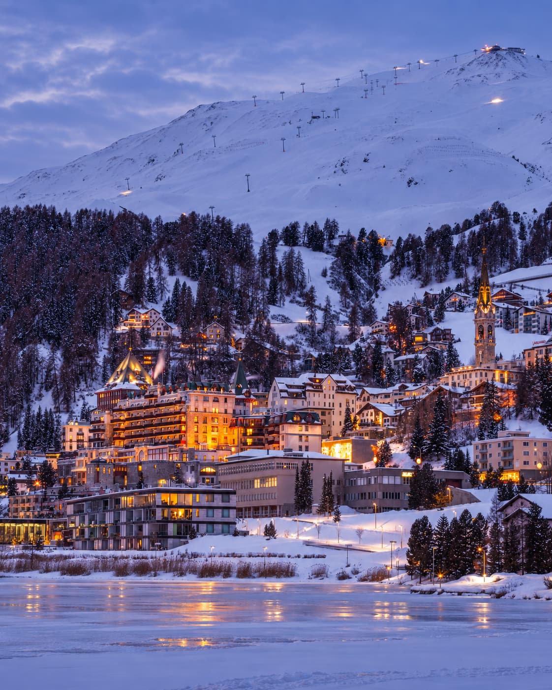 St. Moritz at dusk in winter, amber hotel lights glowing against the snow, frozen lake in the foreground reflecting the lights, ski lifts visible on the mountain above