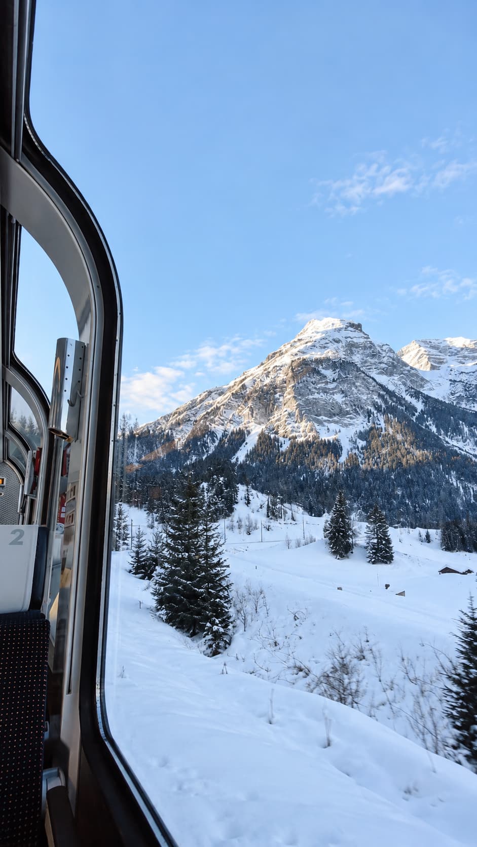 View through a Swiss mountain train window in winter — snowy Alpine peak and valley visible through the glass, travelling Switzerland in winter without skiing